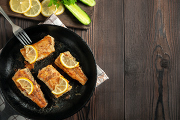 Portioned pieces of fried fish in a black cast-iron pan with fresh cucumbers and herbs. Traditional dinner of a village fisherman, top view, flat lay, copy space