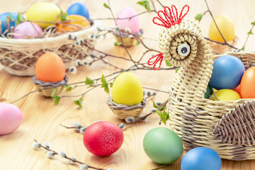 Easter composition - a wicker basket in the form of a chicken with colored eggs on a light wooden table with willow twigs
