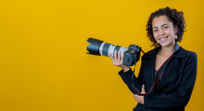 Photo of adorable curly hair woman wearing black clothes holding professional camera isolated over yellow background. Mock up copy space. Photo camera. Professional woman. Photographer's day.