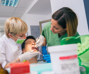 Dentist with the help of her assistant examines the mouth and teeth of a teenage boy patient.