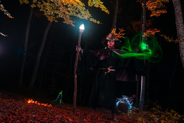 Sorcerer (magician) casting a spell while standing in the magical woods. Holding his magical staff. Magic illumination. Autumn holiday celebration. Mystery and nightmare concept.