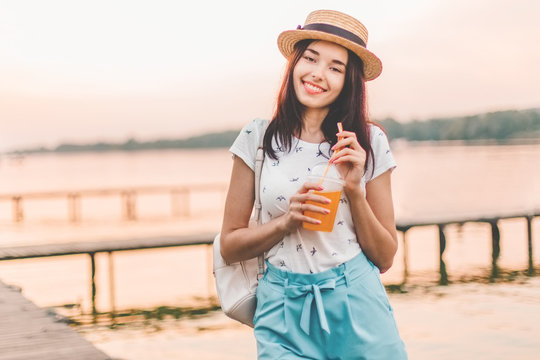 Beautiful young woman walking with orange drink on pier at sunset in summer.