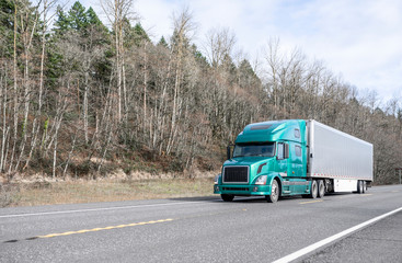 Big rig modern green bonnet semi truck with refrigerator semi trailer running on the road with forest on the hillside