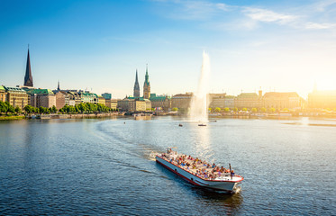 Classic panoramic view of famous Binnenalster (Inner Alster Lake) with fountain, tourist boat and the historic Hanseatic City of Hamburg from Lombardsbridge in golden evening light at sunset, Germany