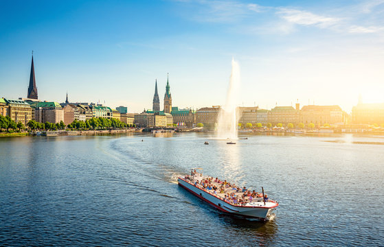 Classic panoramic view of famous Binnenalster (Inner Alster Lake) with fountain, tourist boat and the historic Hanseatic City of Hamburg from Lombardsbridge in golden evening light at sunset, Germany