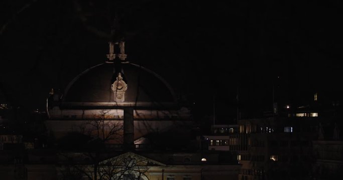 Cinematic shot of illuminated Norwegian National Theatre building in Karl Johan main street with tree and branches in front of camera, at night in Oslo Norway. Close-up/wide shot with parallax motion.