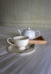 White color ceramic coffee cup with white color teapot on timber trey on white bed sheet