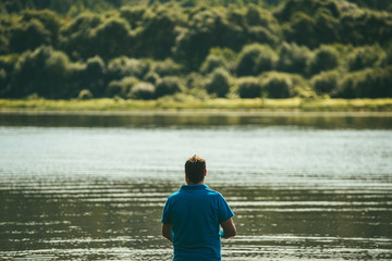 Fototapeta na wymiar a lone man in a blue t-shirt stands on the Bank of the river and catches fish against the background of the forest shore and the water surface. The concept of unity with nature. Back view