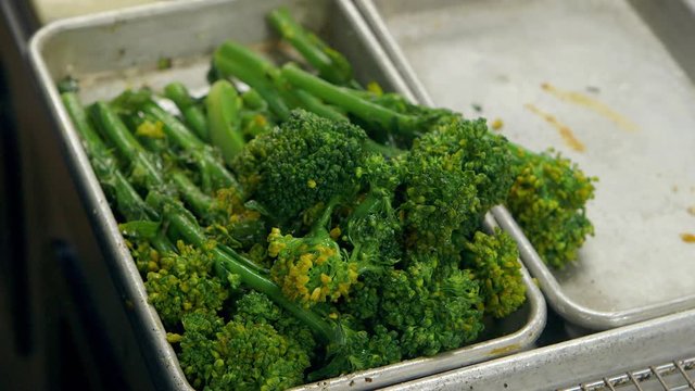 Chef seasons and tosses broccolini in tray in upscale restaurant kitchen
