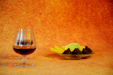 Two glass glasses with cognac and a plate with chocolate candies and cut lemon on a brown abstract...
