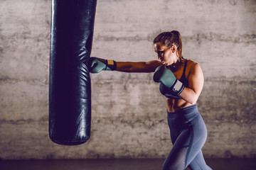 Full length of young dedicated caucasian muscular female boxer in sportswear with ponytail and boxing gloves punching boxing bag while standing in the gym.