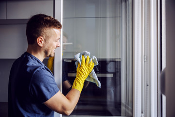 Young bearded smiling positive man with rubber gloves cleaning window with cloth. After rain there is always stains on glass. - obrazy, fototapety, plakaty