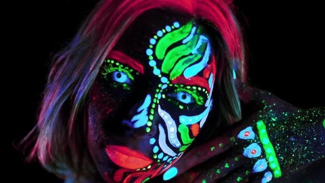 Unusual Body Art with UV Neon Paints. Girl with Neon Lenses in Eyes Looking in to the Camera. Bright Brows, Eyelashes, Lips. Shiny Eyes, Magic Eyes, Bright Eyes, Glows Eye. Macro or Closeup.