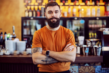 Young handsome bearded tattooed cafe owner standing with arms crossed and looking at camera. - obrazy, fototapety, plakaty