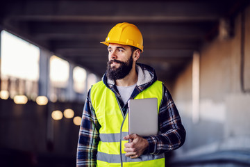 Portrait of highly motivated caucasian hardworking smiling bearded supervisor with helmet on head in vest and with laptop in hands posing on construction site.
