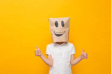Cute little school boy standing with a paper bag on his head - happy face on yellow background, hppy emotion concept