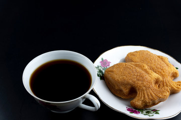 Traditional Malaysian cake called "kuih bahulu or baulu" in fish shape, in flowery plate with a cup of coffee over dark background.