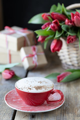 Red cup of coffee close-up, a bouquet of red tulip and gift boxes on wooden background