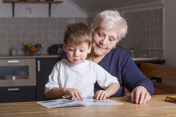 Portrait of beautiful mature woman (80 years old) with her great-grandson at home, reading educational book together