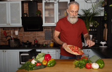 Picture of mature handsome bearded man standing at the kitchen and cooking salad