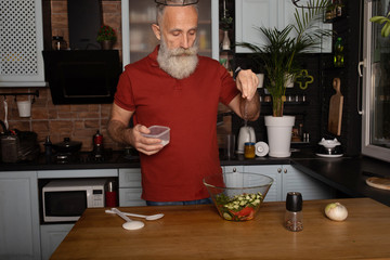 Picture of mature handsome bearded man standing at the kitchen and cooking salad