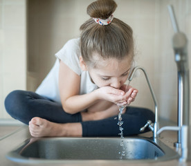 Caucasian cute girl is drinking from water tap or faucet in kitchen at home. Hydration. Lifestyle....