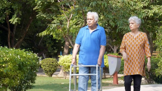 Retired old man doing a morning walk using a walker with his wife in a garden. Senior white-haired husband and wife happily talking while walking together on a sunny day in a park - healthy lifesty...
