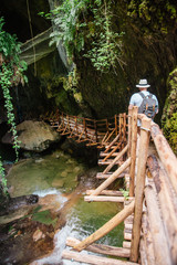 young strong man in white hat with backpack at cave entrance. hiking concept. creek with waterfall