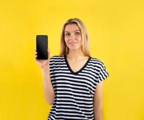 Joyful woman demonstrating smartphone blank display screen to camera on yellow background. Copy...