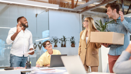 Moving to a new office. Young handsome man in casual wear holding box with things and waving to his new coworkers while standing in the modern office