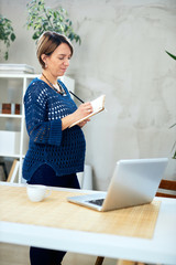 Attractive pregnant businesswoman taking notes in modern office interior.