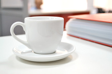 Coffee break. White cup for tea or coffee on a white office desk near a red file folder and hardcover. The cup is empty free in the workspace. Against the background of blurred office space.