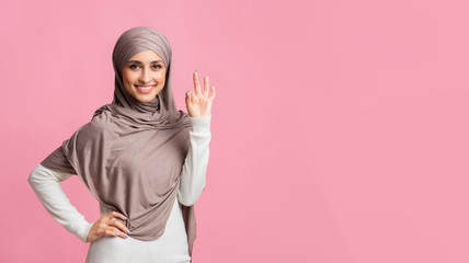 Smiling arabic girl in headscarf showing ok sign on pink background
