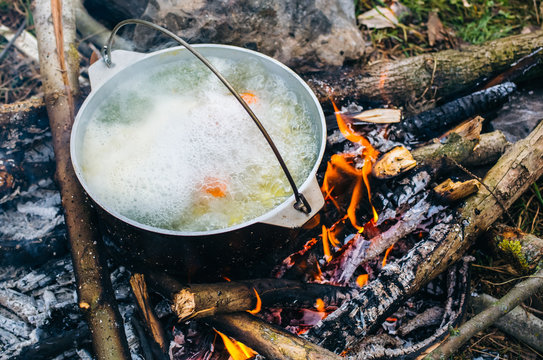 Old vintage pot in black soot is standing on the open fire in the wood. Natural summer holidays photo. Fish broth cooked in the forest. Vegetarian carrots and potatoes vegetable soup. Camp