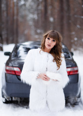 A beautiful pretty cheerful funny young girl with curly hair and a snow white smile in a white fur coat and a warm hat looks out of the car window against the background of a winter forest