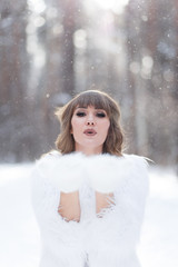 A beautiful pretty young girl with curly hair and a snow-white smile in a white warm fur coat is walking and fooling around in the winter forest against the background of snow and trees, enjoying