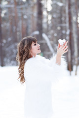 winter portrait of a beautiful attractive young cheerful curly haired girl in a white fur coat with a snow heart in her hands on the background of a winter forest