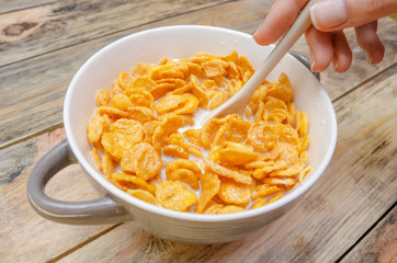 girl's hand with a white spoon mixes corn flakes in a bowl with milk on a wooden background