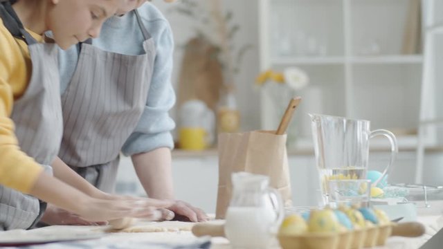 Cheerful grandma in apron helping little granddaughter with rolling out pastry dough while cooking together at kitchen table