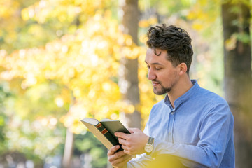 Handsome young man reading a book in a park. Portrait of a young man with a denim jacket and blue shirt reading a book outside. A guy reading a book in a park