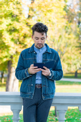 Handsome young man reading an ebook in a park. Portrait of a young man with a denim jacket and blue shirt reading an e-book outside. A guy reading an ebook in a park