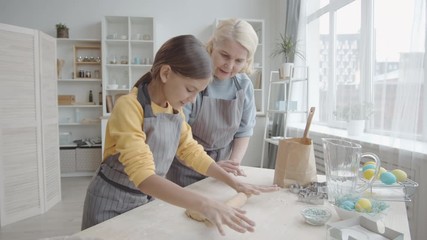 Zoom in shot of beautiful girl in apron rolling out pastry dough with pin on kitchen table while baking under control of helpful grandmother