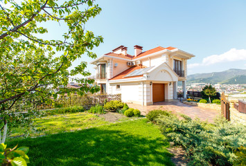 The facade of a classic Mediterranean two-storey cottage in the spring. In front of the cottage there is a green lawn and flowering trees. Mountains in the background.