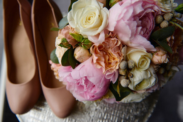 Bridal bouquet of pink peonies, coral roses and greenery with satin ribbon and beige women shoes on round table, copy space. Wedding concept
