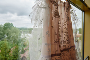 Beautiful beige embroidered wedding dress hanging on hanger against window in the room, copy space, close up. Bridal morning preparations