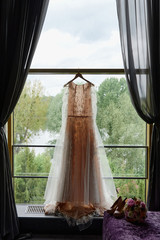 Beautiful beige wedding dress hanging on hanger against window in hotel room, copy space. Bridal bouquet and women's shoes standing on sofa
