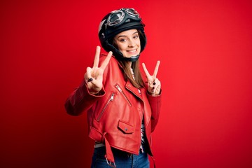 Young beautiful brunette motocyclist woman wearing motorcycle helmet and red jacket smiling looking to the camera showing fingers doing victory sign. Number two.