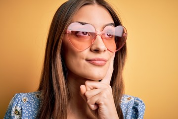 Young beautiful brunette woman wearing funny sunglasses with heart over yellow background serious face thinking about question, very confused idea