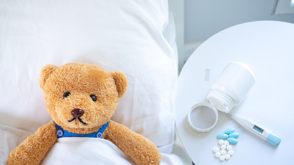 The teddy bear is sick in the bed next to him. There is medication, thermometer waiting to be treated.