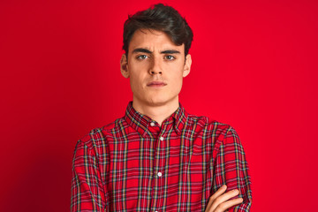 Teenager boy wearing red shirt standing over isolated background skeptic and nervous, disapproving...